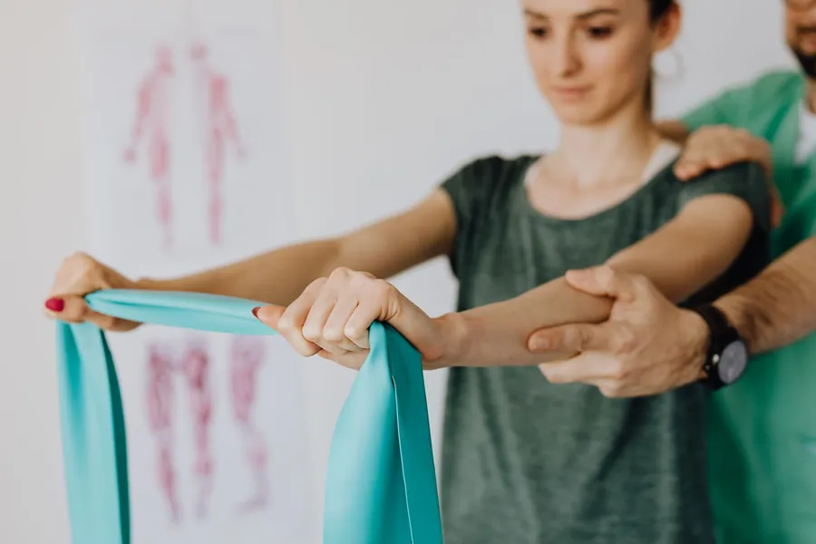 A physical therapist supports a patient's arm as she pulls a resistance band