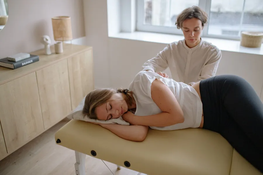 A patient rests on her side on a treatment table while a clinician works on her low back in a calm room with wood furniture
