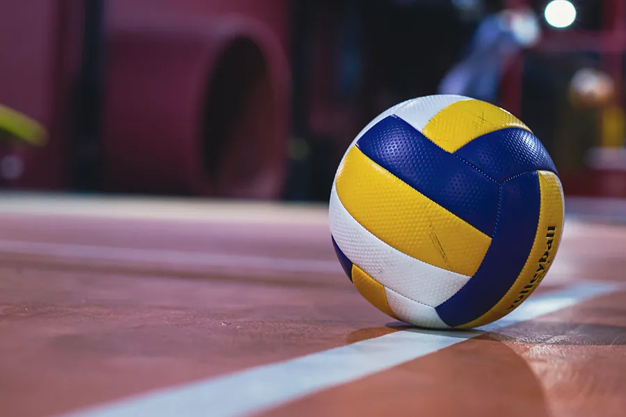 A yellow, blue and white volleyball resting on a white line of an indoor court under arena lights