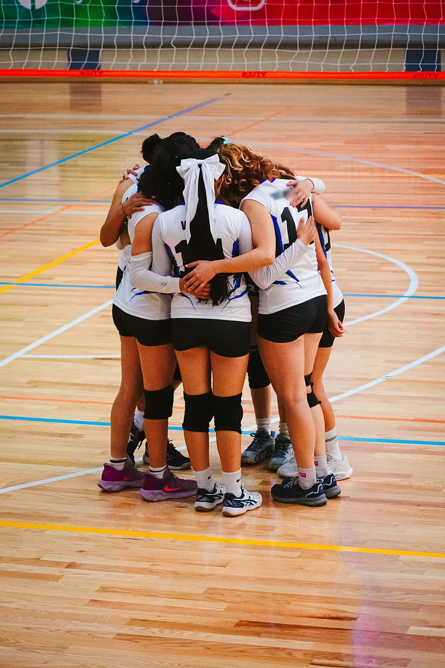 Volleyball players in white jerseys and knee pads in a tight huddle on a gym floor, seen from behind