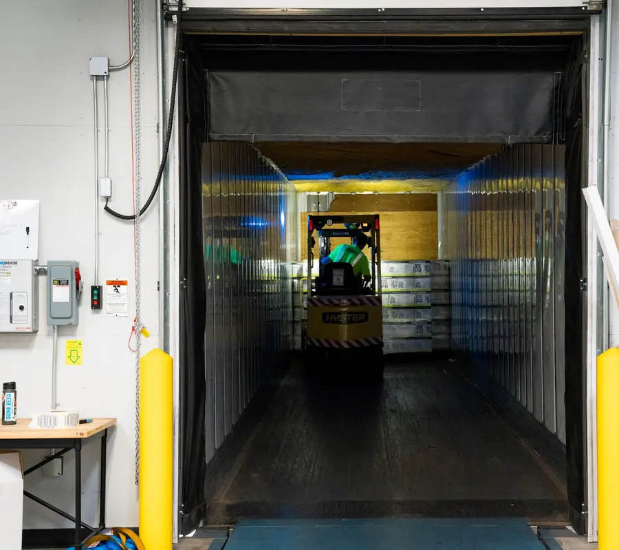 Looking from a loading dock into an open trailer while a forklift loads pallets inside.