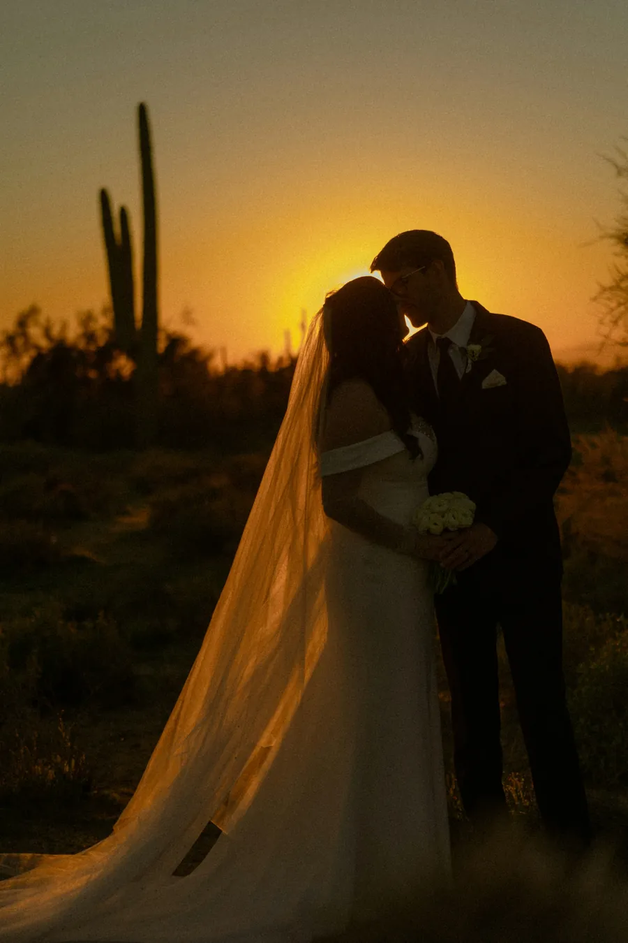 A couple silhouetted against an orange desert sunset beside a tall saguaro cactus, the bride's veil trailing behind her
