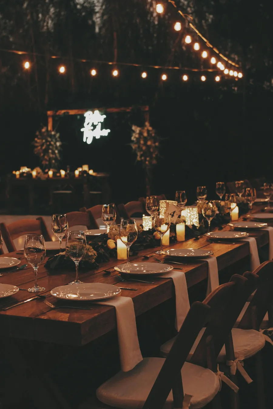 Long wooden reception tables set with candles, glassware and napkins beneath strings of glowing lights