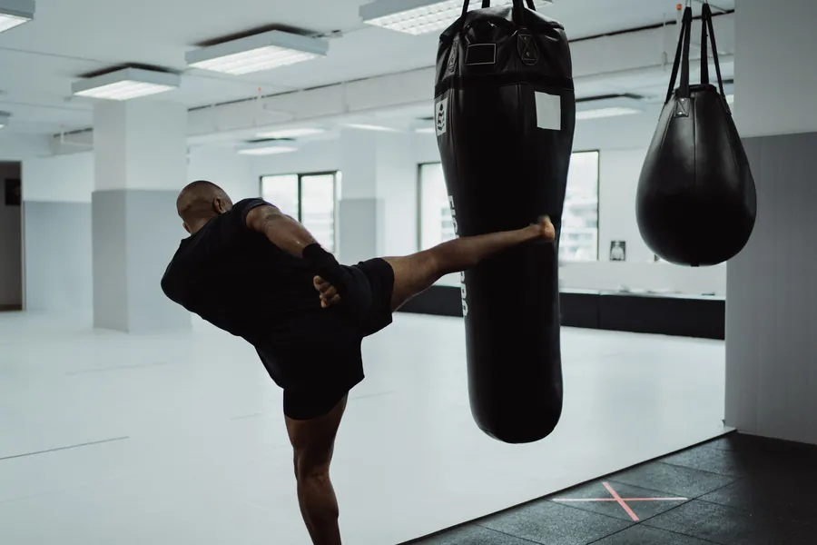 A man throws a high kick into a heavy bag in a bright, open studio