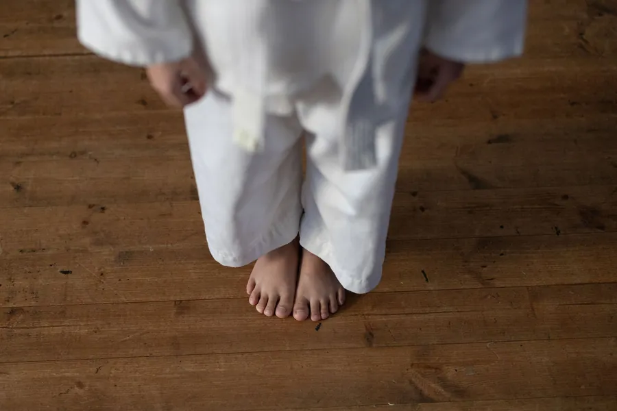 A child in a white gi stands barefoot on a wooden studio floor