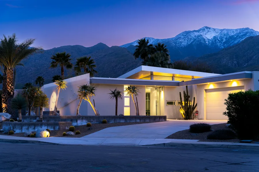 A single-story desert modern home at dusk, its windows and floating roof lit warm, with palms and snow-capped mountains behind