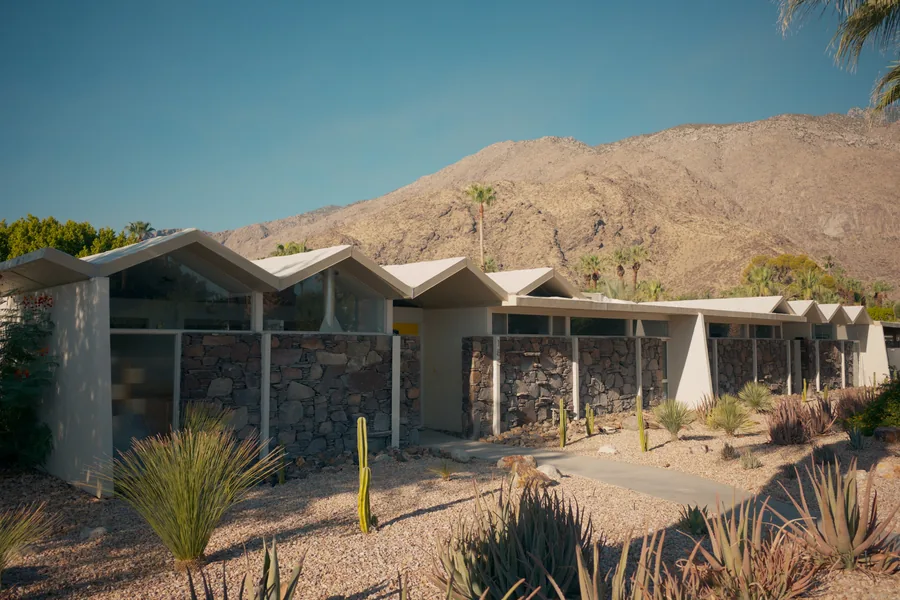 A long, low modern home with stone walls and folded roofs in a gravel and cactus garden below desert mountains