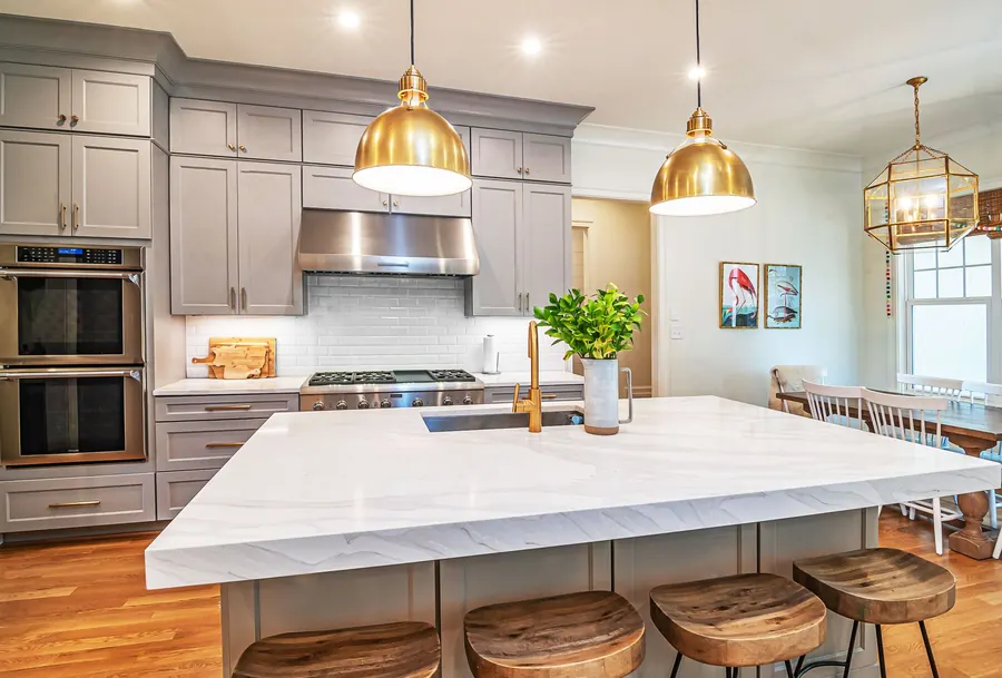 A remodeled kitchen with a large marble island, wood stools and brass pendant lights