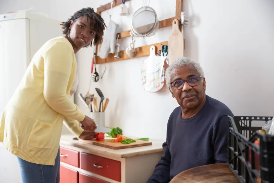 A caregiver chops vegetables in a home kitchen while an older man sits nearby and smiles