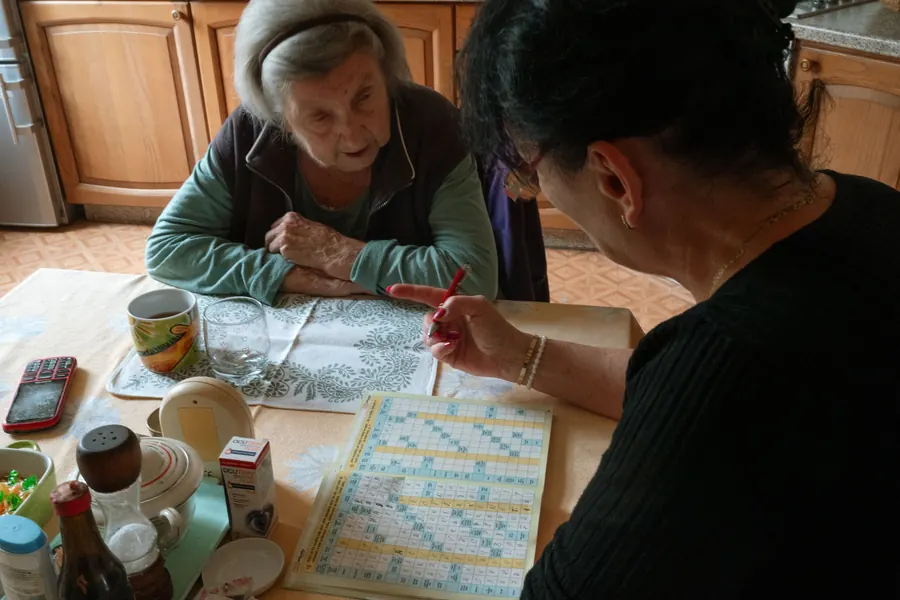 An older woman and her caregiver work on a crossword puzzle together at a kitchen table