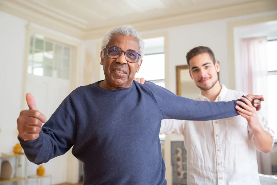 A young caregiver supports a smiling older man through a gentle arm stretch in his living room