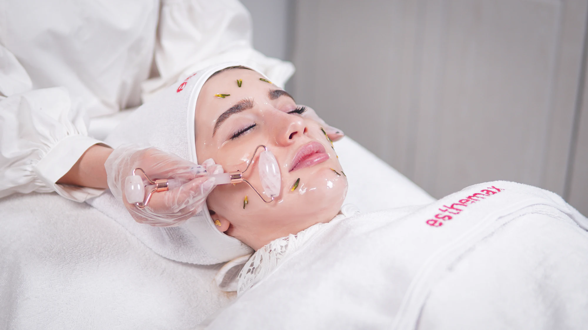 A guest wrapped in a soft towel rests while an esthetician glides a rose-quartz roller along her cheek