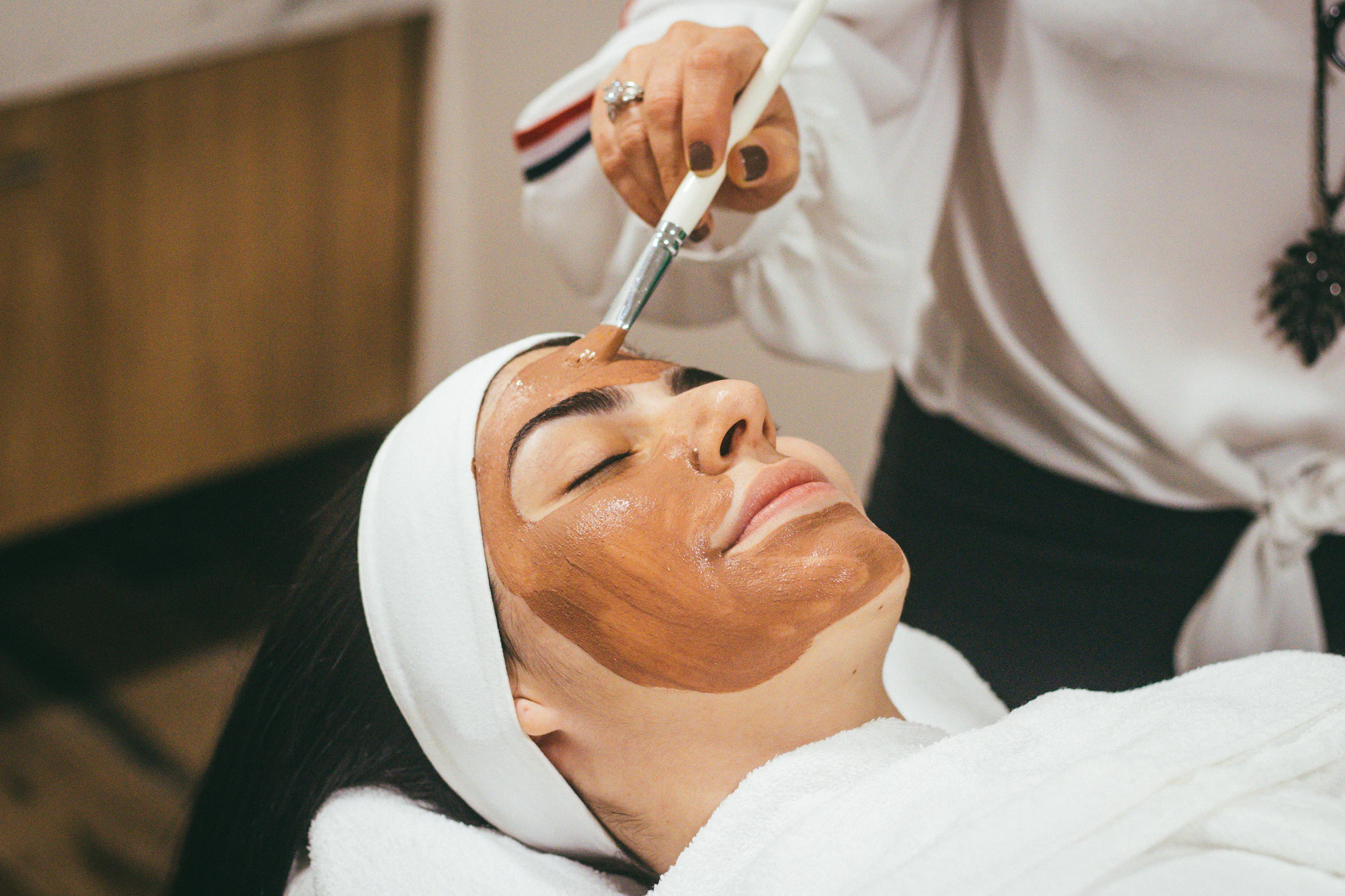 An esthetician brushes a warm clay mask onto a relaxed guest's face during a facial
