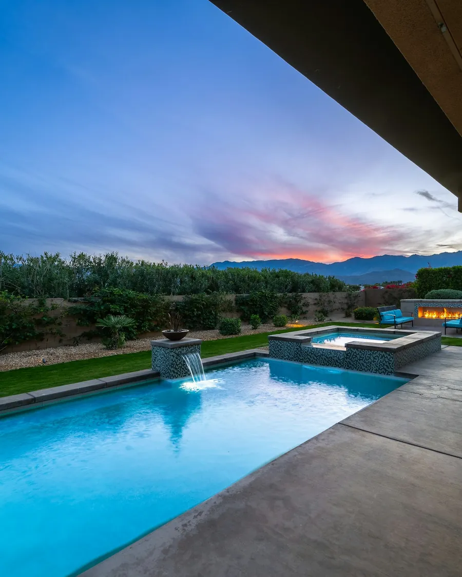 A backyard pool and raised spa at dusk, with a sheer-descent water feature, a lawn, and desert mountains under a pink sky