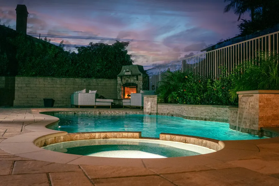A lit pool with a round raised spa and stone deck at dusk