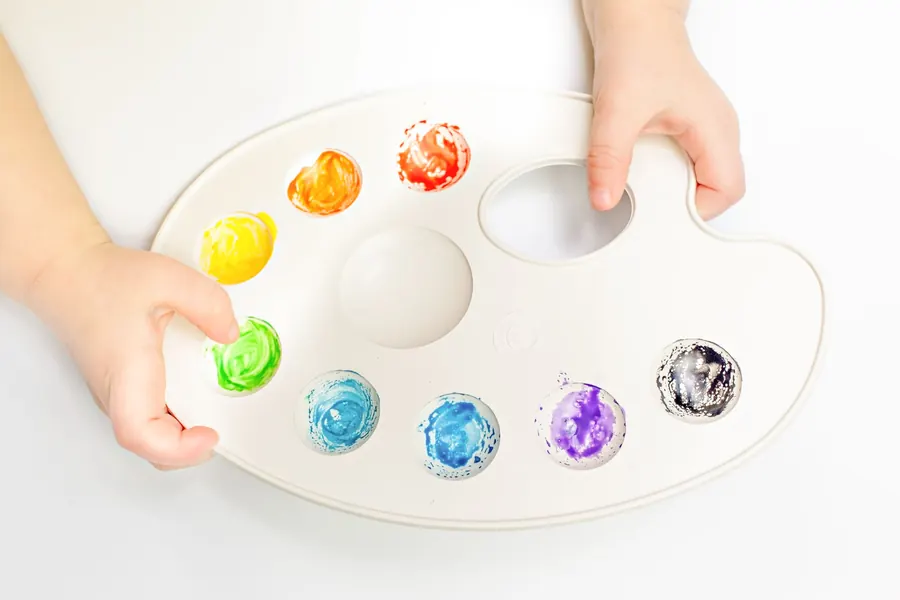 A small child's hands holding a white paint palette with bright colors