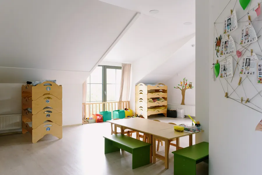 A bright classroom with low wooden shelves, green benches and a long work table